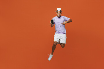 Full body traveler black man wear purple t-shirt hat hold mobile phone jump isolated on plain orange color background. Tourist travel abroad in spare time rest getaway Air flight trip journey concept