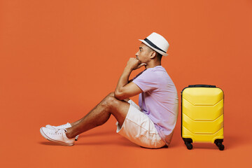 Full body traveler black man wear purple t-shirt hat sit near bag hold head isolated on plain orange color background Tourist travel abroad in spare time rest getaway Air flight trip journey concept.
