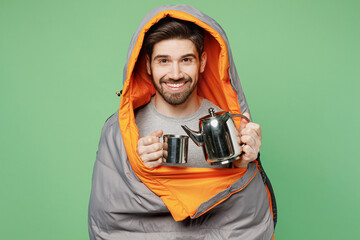 Young fun traveler white man in sleeping bag hold cup kettle pours tea isolated on plain green...