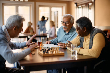 Senior man talks to his friends who are playing chess at nursing home. - obrazy, fototapety, plakaty