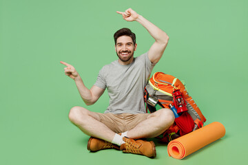 Full body young traveler white man near backpack mat sit on ground point finger aside on area...