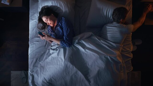 Top View: Young Woman Uses Smartphone in Bed at Night When Her Female Partner Trying to Fall Asleep Beside. Couple Fight, Argue. Addictive World of Social Media, Doom Scrolling, Fake News.