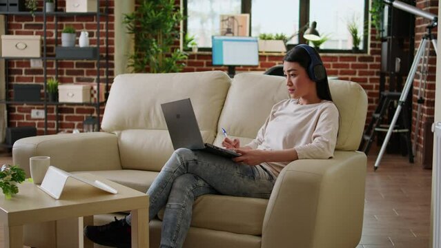 Focused working woman wearing wireless headphones taking notes while doing remote work at home sitting on sofa. Serious looking student attending online class on modern laptop.