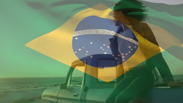 Animation of brazilian flag waving over african american woman sitting on pick-up truck at beach