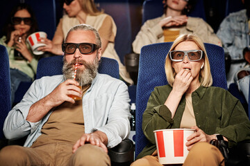 Portrait of mature Caucasian man and woman wearing 3D glasses watching movie, eating popcorn and sipping fizzy drink at cinema