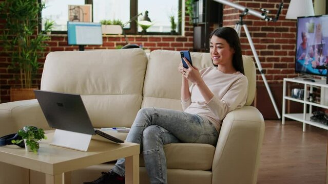 Joyful young adult person texting with friend while sitting on sofa inside apartment. Smiling heartily woman watching funny content on smartphone while drinking coffee inside living room on couch.