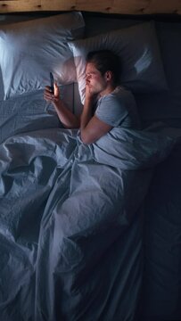 Vertical Screen: Caucasian Man Uses Smartphone in Bed at Home at Night. Handsome Guy Browsing Social Media, Reading News, Doing Online Shopping, Chatting with Friends at Night. Holding Mobile Phone