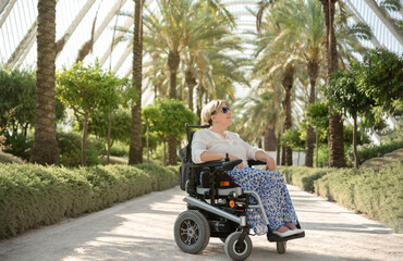 a smiling woman sitting in the electric wheelchair with disability enjoys a sunny day in the garden of the city park
