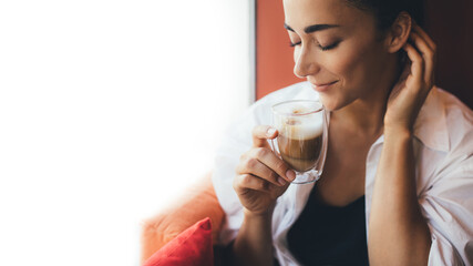 Young woman drinking coffee cappuccino sitting near window.