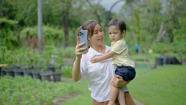 Family recreation and weekend leisure in park. Asian mother makes a selfie with her little son in Green Summer Park, Mom taking photos using mobile phone, They gladly smile at the camera. 