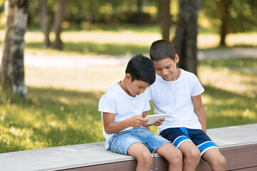 Two asian boys, brothers, using together a digital tablet. Children sitting outdoor in public park in summer, selective focus.