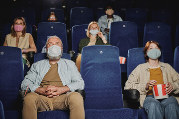 Group of people wearing masks, keeping distance from each other watching movie at cinema during...