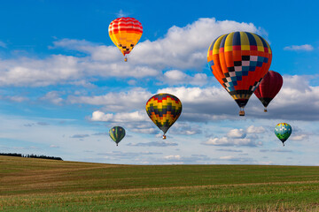Multicolored hot air balloons fly in blue sky with white clouds over green field.