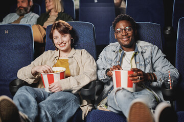 Portrait of stylish young Black man and Caucasian wiman sitting relaxed with popcorn on lap at...