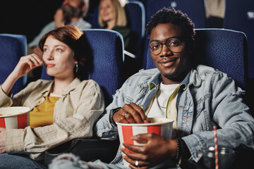 Selective focus shot of joyful young African American man wearing casual clothes sitting with his girlfriend at cinema smiling at camera