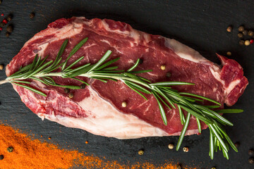 Raw appetizing steak with seasonings on a black stone serving board. Rosemary, paprika, black peppercorns. Close-up. Top view.