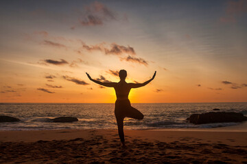 Fototapeta na wymiar Silhouette slim woman does yoga position arms raised on tropical sea coast or ocean beach outdoors at sunset