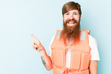 Young caucasian man wearing life jacket isolated on blue background smiling and pointing aside,...