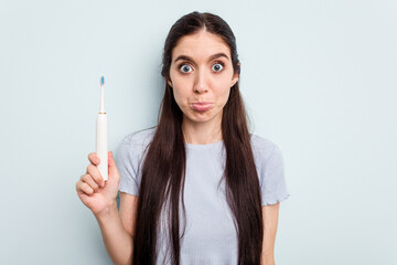Young caucasian woman holding a electric toothbrush isolated on blue background shrugs shoulders...