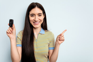 Young caucasian woman holding car keys isolated on blue background smiling and pointing aside, showing something at blank space.