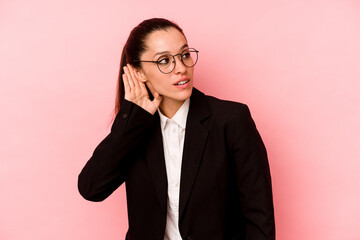 Young business caucasian woman isolated on pink background trying to listening a gossip.