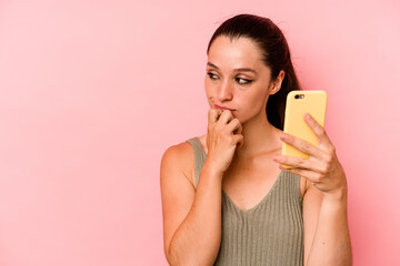 Young caucasian woman holding mobile phone isolated on pink background looking sideways with doubtful and skeptical expression.