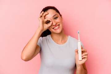 Young caucasian woman holding a electric toothbrush isolated on pink background excited keeping ok gesture on eye.