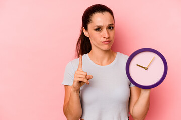 Young caucasian woman holding a clock isolated on pink background showing number one with finger.