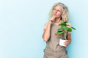 Middle age gardener woman holding a plant isolated on blue background looking sideways with...