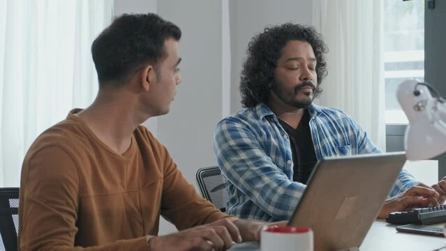 Waist up slowmo of two male Indian programmers talking while working on computers at desk in office