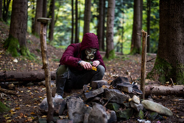 a boy in a gas mask and a cherry jacket sits in the middle of the forest and starts a fire. nuclear war concept