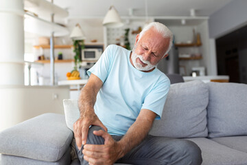 The older man is sitting on the couch at home, has pain in the knee joint, holding his leg, osteoarthritis concept. - obrazy, fototapety, plakaty