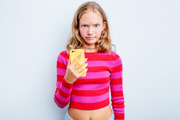 Caucasian teen girl holding mobile phone isolated on blue background shrugs shoulders and open eyes confused.