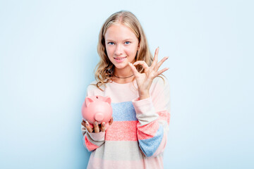 Caucasian teen girl holding a piggybank isolated on blue background cheerful and confident showing ok gesture.
