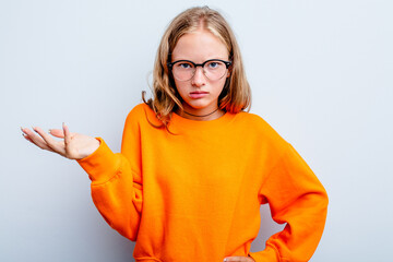 Caucasian teen girl isolated on blue background doubting and shrugging shoulders in questioning...