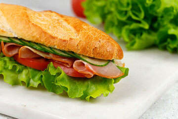 Baguette sandwich with ham, tomato, lettuce, cucumber on a marble board and grey background. Close up Sandwich