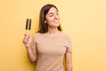 Young caucasian woman holding a brush hair isolated on yellow background looks aside smiling, cheerful and pleasant.