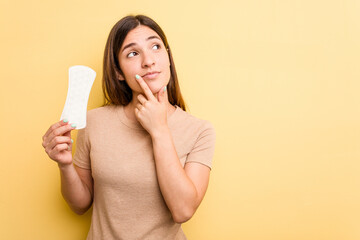 Young caucasian woman holding a sanitary napkin isolated on yellow background looking sideways with doubtful and skeptical expression. - obrazy, fototapety, plakaty