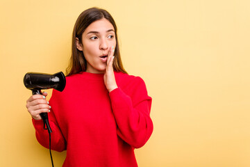 Young caucasian woman holding hairdryer isolated on yellow background is saying a secret hot braking news and looking aside