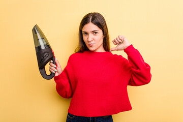 Young caucasian woman holding a hand vacuum cleaner isolated on yellow background feels proud and...