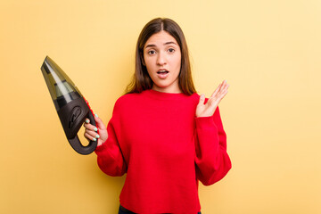 Young caucasian woman holding a hand vacuum cleaner isolated on yellow background surprised and shocked.