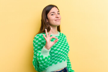 Young caucasian woman isolated on yellow background winks an eye and holds an okay gesture with hand.