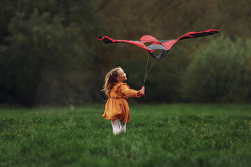 Having fun. Little girl is running with red colored kite outdoors. Playful mood - Powered by Adobe