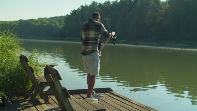 Skillful handsome black male fisherman standing on wooden jetty, casting spin fishing rod while enjoying leisure and fishing on lake in rays of warm summer sunrise.