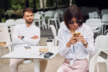 Couple sitting together outdoors. Woman using smartphone, man have his arms crossed
