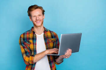 Photo of clever handsome cheerful guy with ginger hair dressed plaid shirt typing code at remote job isolated on blue color background