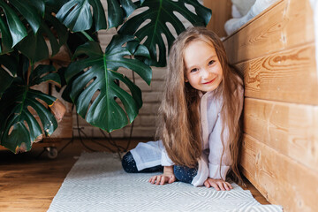 Little beautiful girl with long hair sits on a wooden floor near large leaves of a monstera flower.
