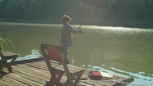 Positive skillful adorable preadolescent Middle Eastern boy standing on wooden pier, casting spin fishing rod while enjoying leisure and freshwater fishing on lake at daybreak