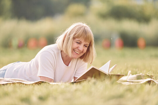 Laying down and reading book. Senior woman having nice weekend outdoors on the field at sunny day
