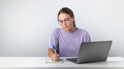 The girl sits at the laptop and makes notes in the notebook, study, work